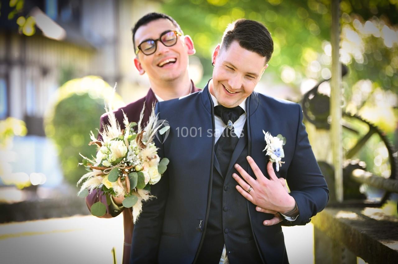 Deux hommes souriants en tenue de mariage, l'un tenant un bouquet, dans un cadre extérieur ensoleillé.