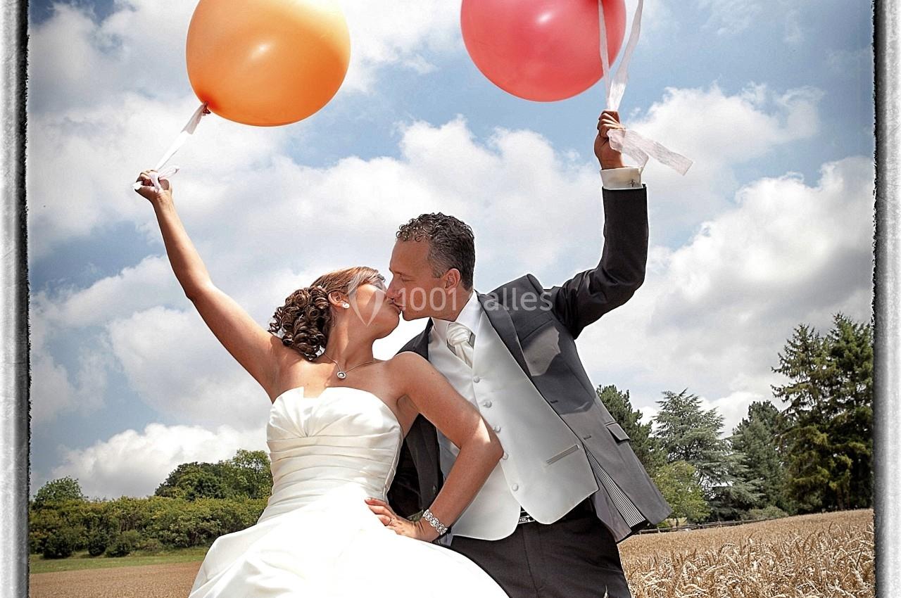 Un couple en tenue de mariage s'embrasse dans un champ de blé, tenant des ballons colorés sous un ciel bleu.