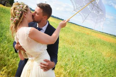 Un couple en tenue de mariage s'embrasse dans un champ de blé, tenant des ballons colorés sous un ciel bleu.