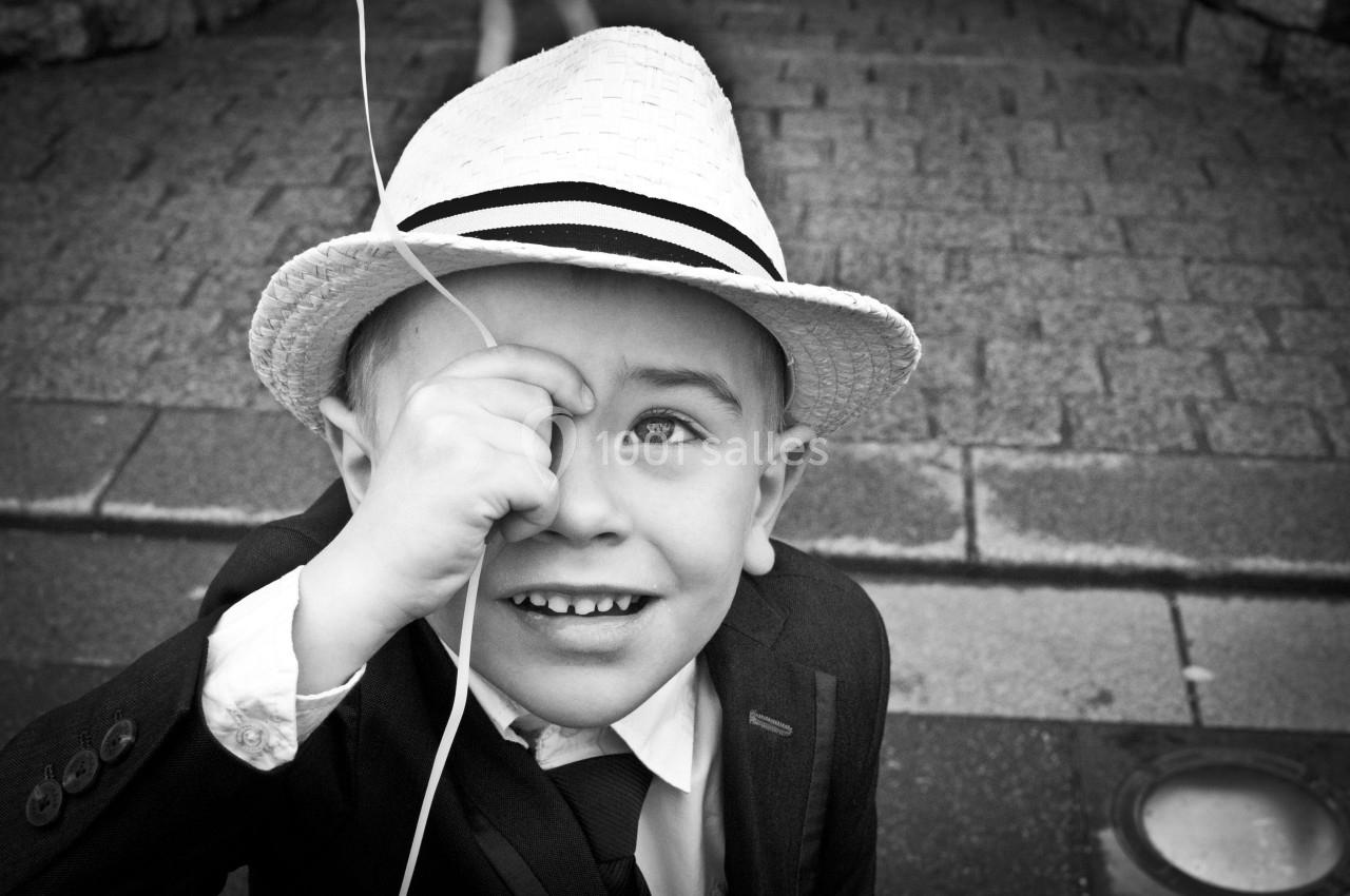Un enfant souriant en costume et chapeau blanc tient un ballon devant son œil, sur un trottoir pavé.