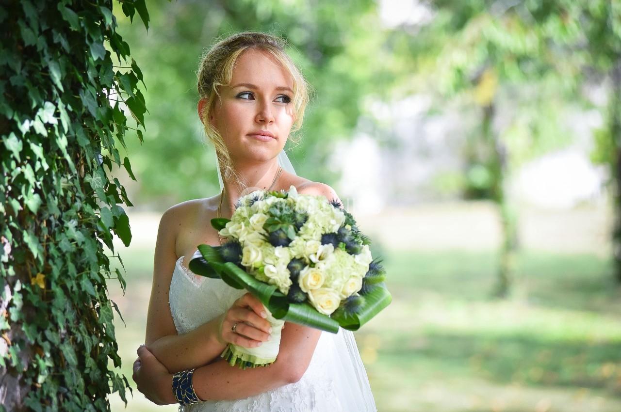 Une mariée en robe blanche tient un bouquet de fleurs blanches et vertes dans un jardin verdoyant.