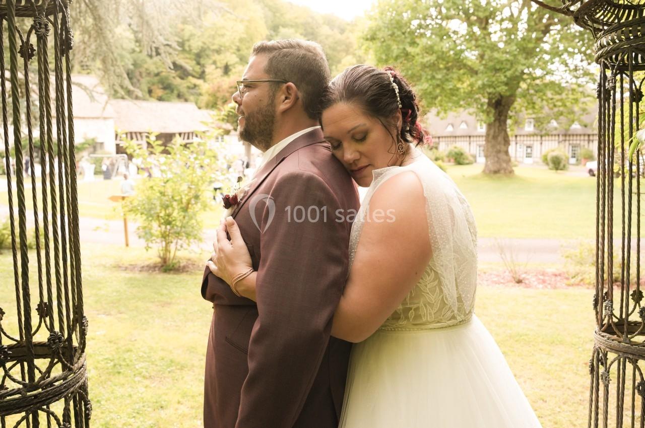 Une femme en robe de mariée enlace un homme en costume dans un jardin verdoyant près d'un bâtiment ancien.