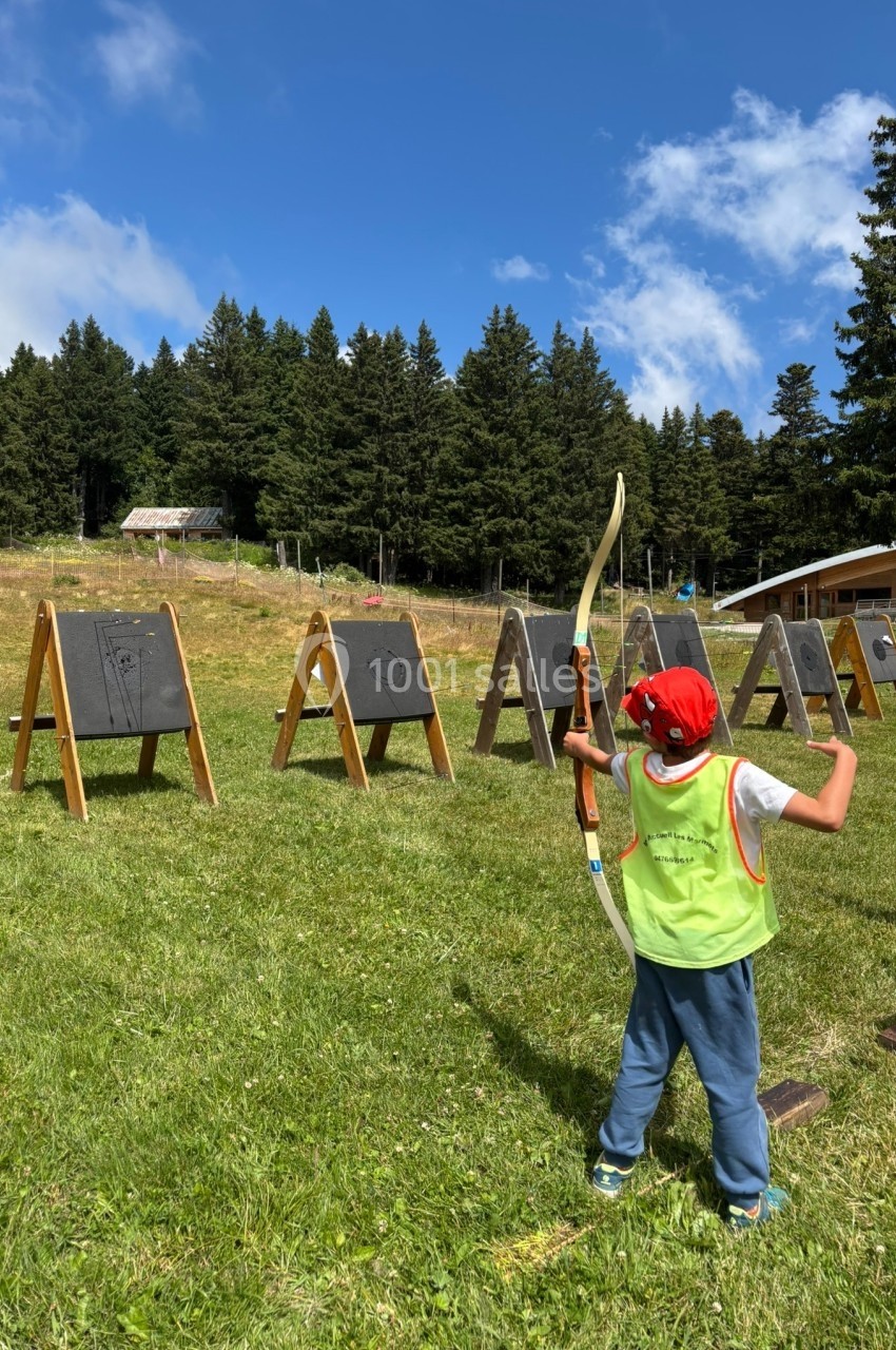 Un enfant en tenue de tir à l'arc vise une cible en plein air sur une pelouse, entouré de forêts et de ciel bleu.