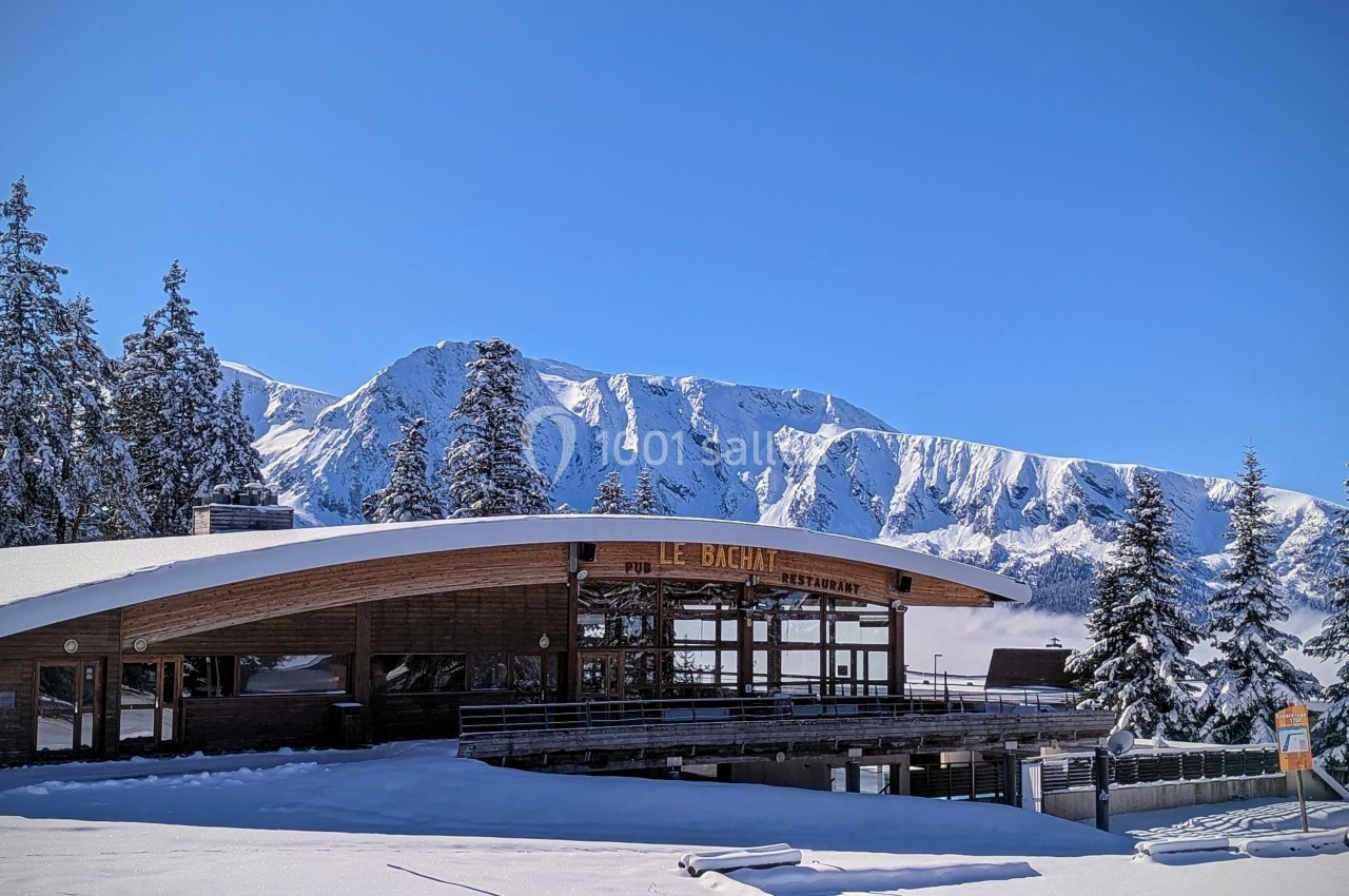 Chalet en bois avec terrasse enneigée, entouré de sapins, devant une chaîne de montagnes sous un ciel bleu.