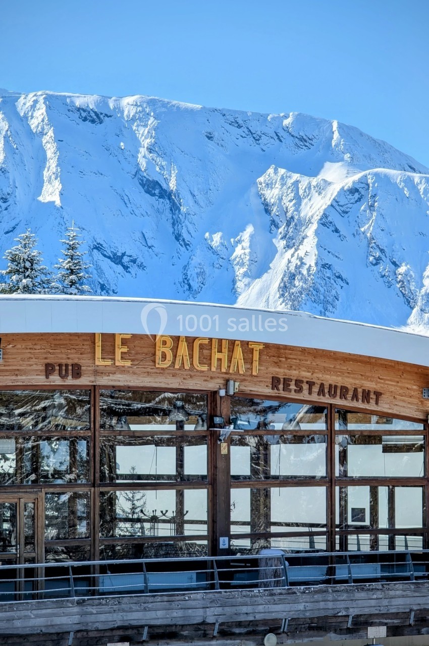 Façade en bois du restaurant ’Le Bachat’ avec des montagnes enneigées en arrière-plan sous un ciel bleu.