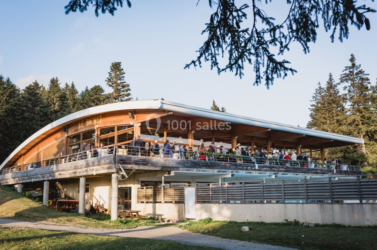 Bâtiment moderne en bois avec terrasse bondée, entouré de verdure et de sapins sous un ciel dégagé.