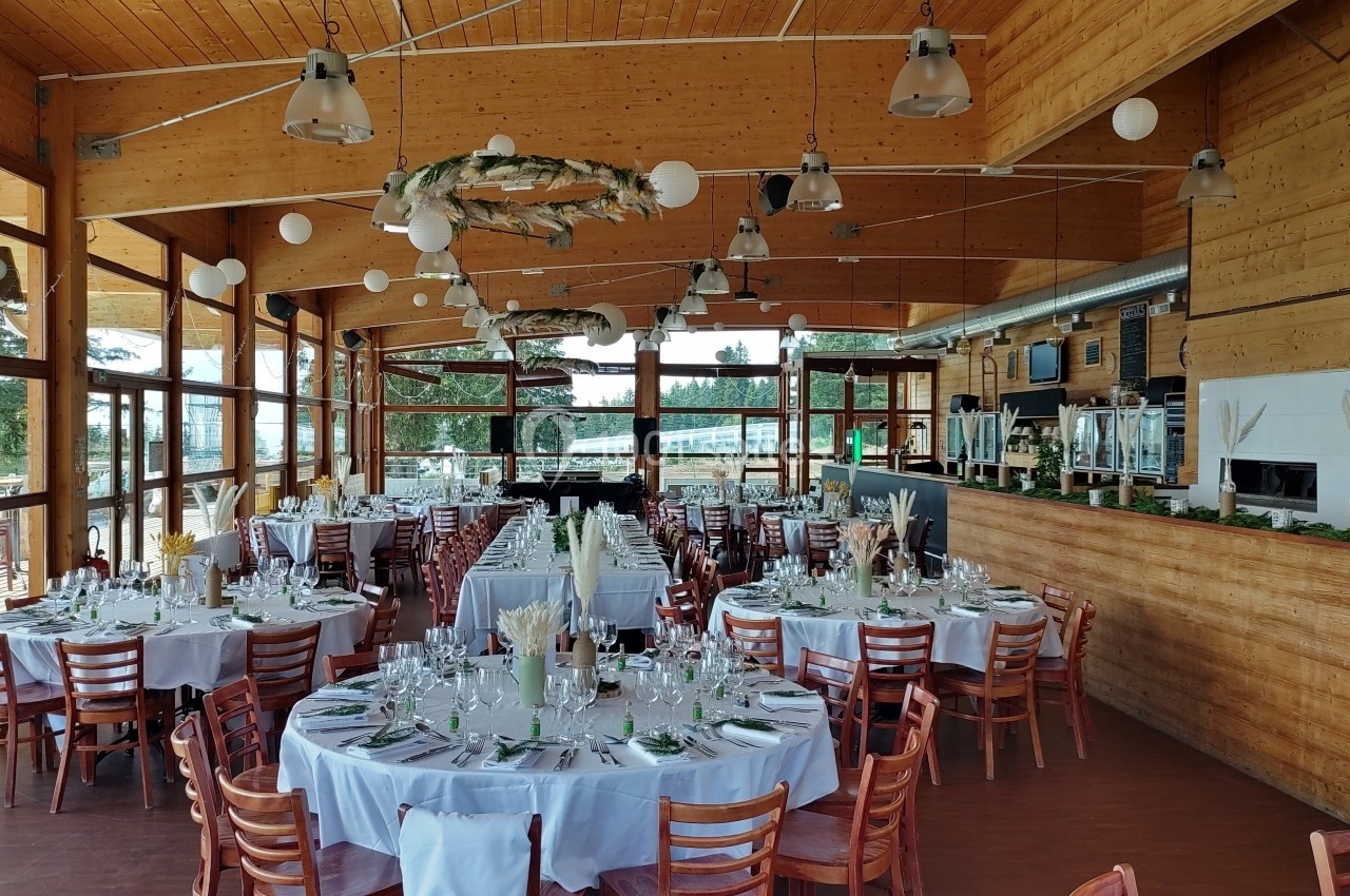 Salle de réception en bois avec tables rondes dressées pour un événement, décorée de guirlandes et de luminaires suspendus.