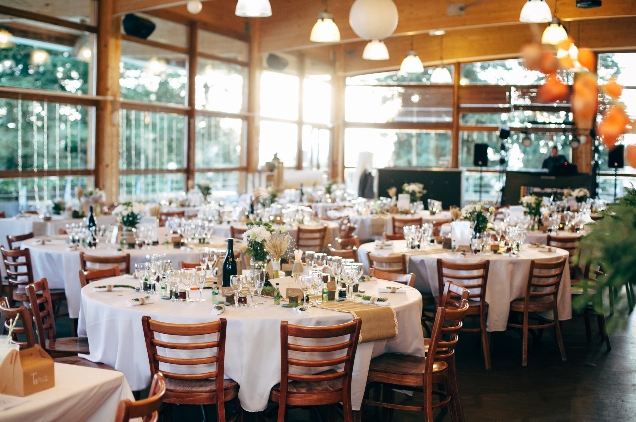 Salle de réception lumineuse avec tables rondes dressées, nappes blanches, chaises en bois et décoration florale.