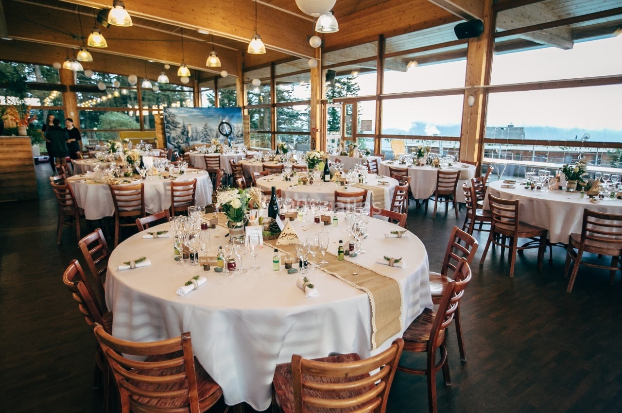 Salle de réception lumineuse avec tables rondes dressées pour un événement, entourées de chaises en bois.