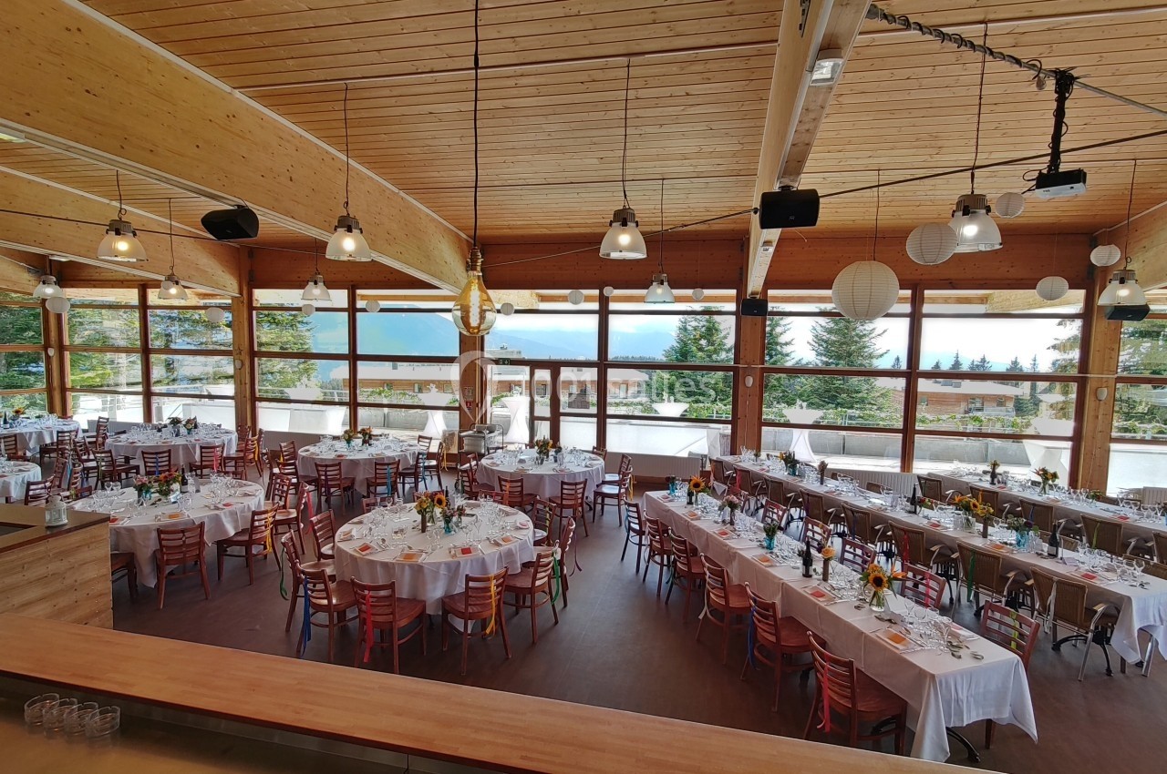 Salle de réception lumineuse avec de grandes baies vitrées, tables dressées pour un événement et vue sur un paysage boisé.