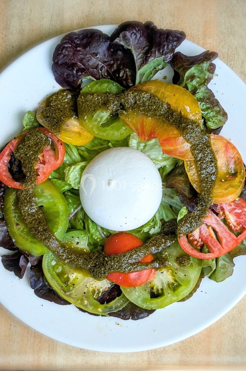Assiette de burrata entourée de tranches de tomates colorées, salade verte et pesto, servie sur une table en bois.