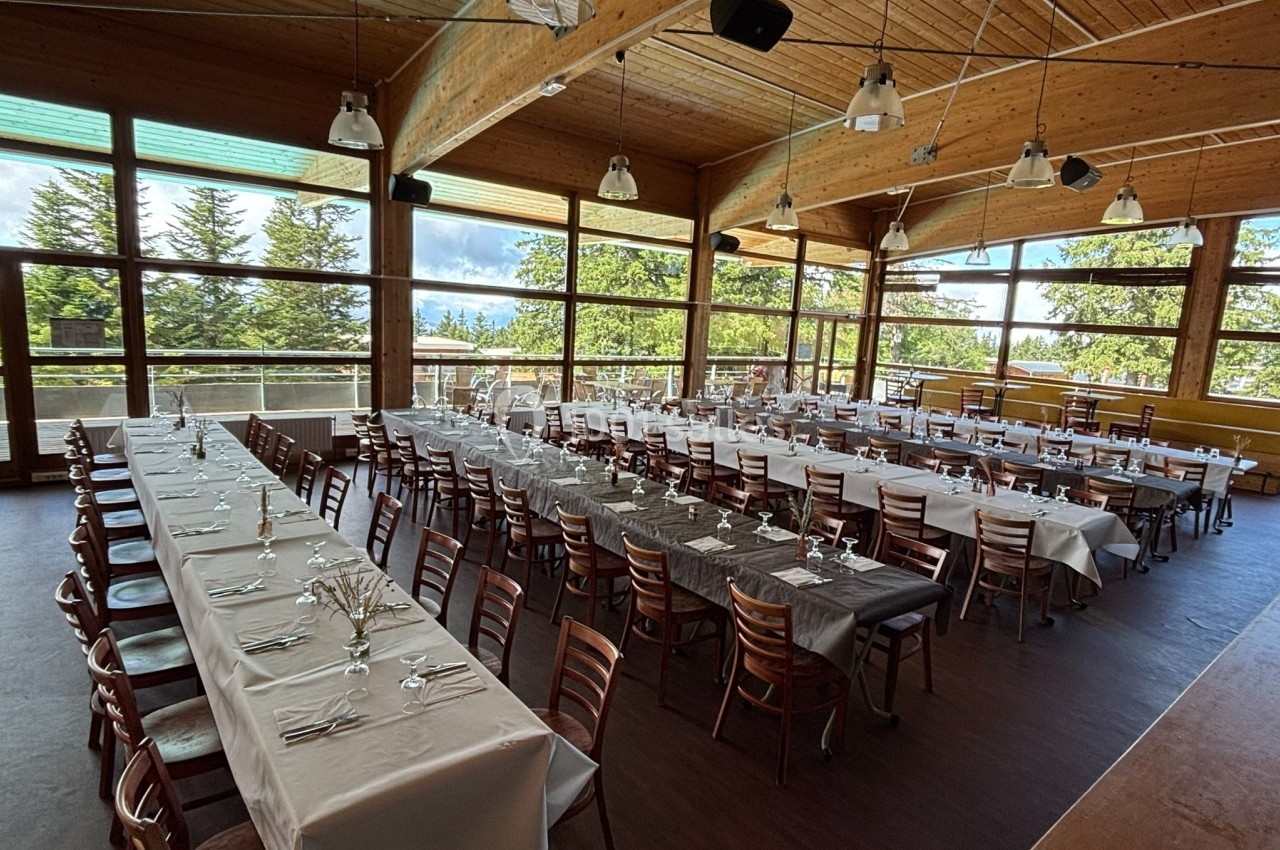 Salle lumineuse avec de grandes baies vitrées, tables longues dressées avec nappes blanches et grises, chaises en bois.