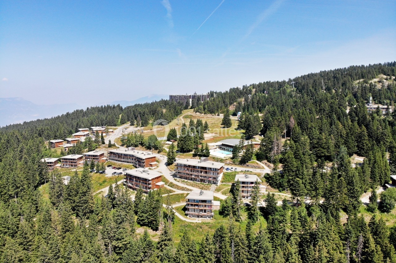 Vue aérienne d'une station de montagne avec des bâtiments entourés de forêts et des collines verdoyantes.