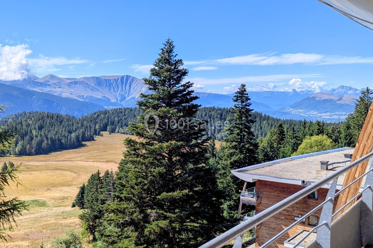 Vue depuis un balcon sur une vallée entourée de montagnes, avec des sapins et un bâtiment en premier plan.