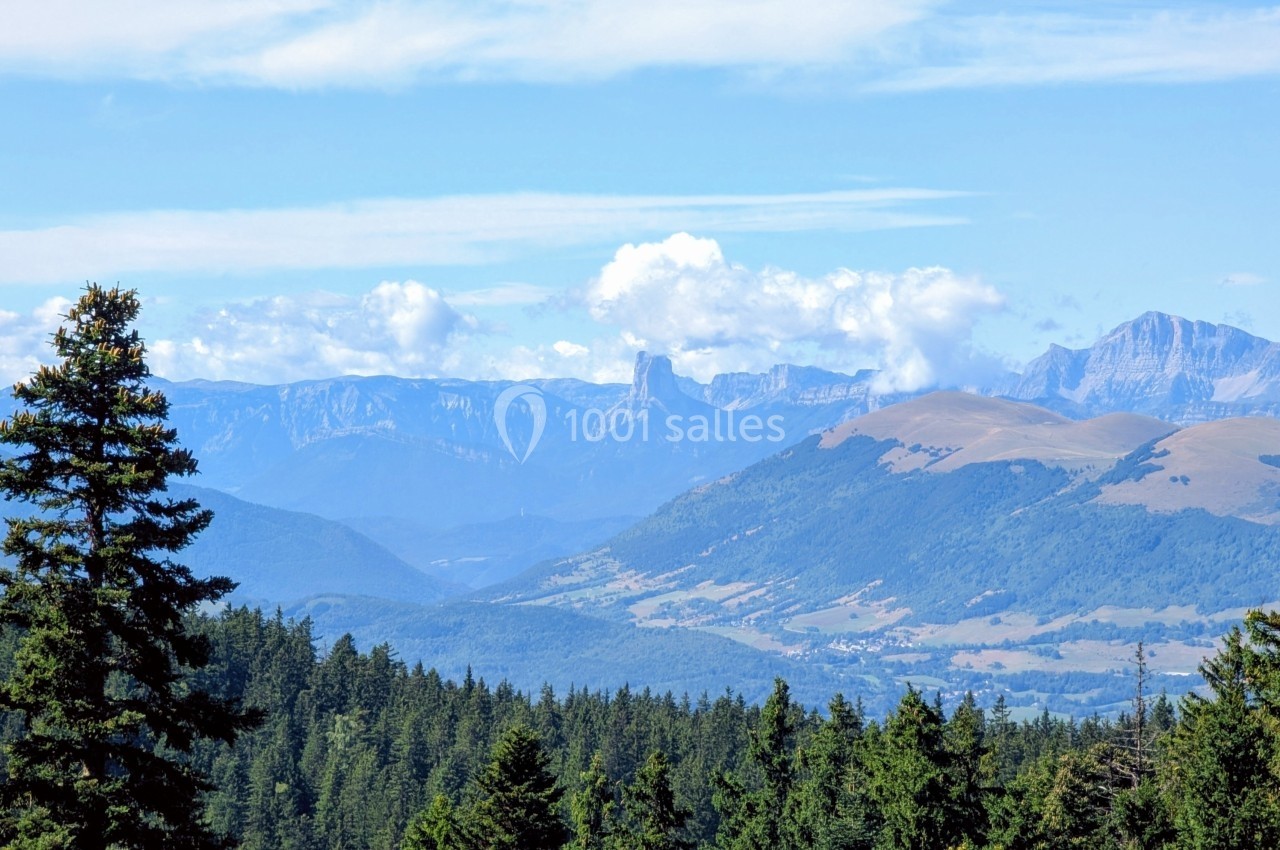 Paysage de montagne avec une vallée boisée, des sommets au loin et un ciel partiellement nuageux.