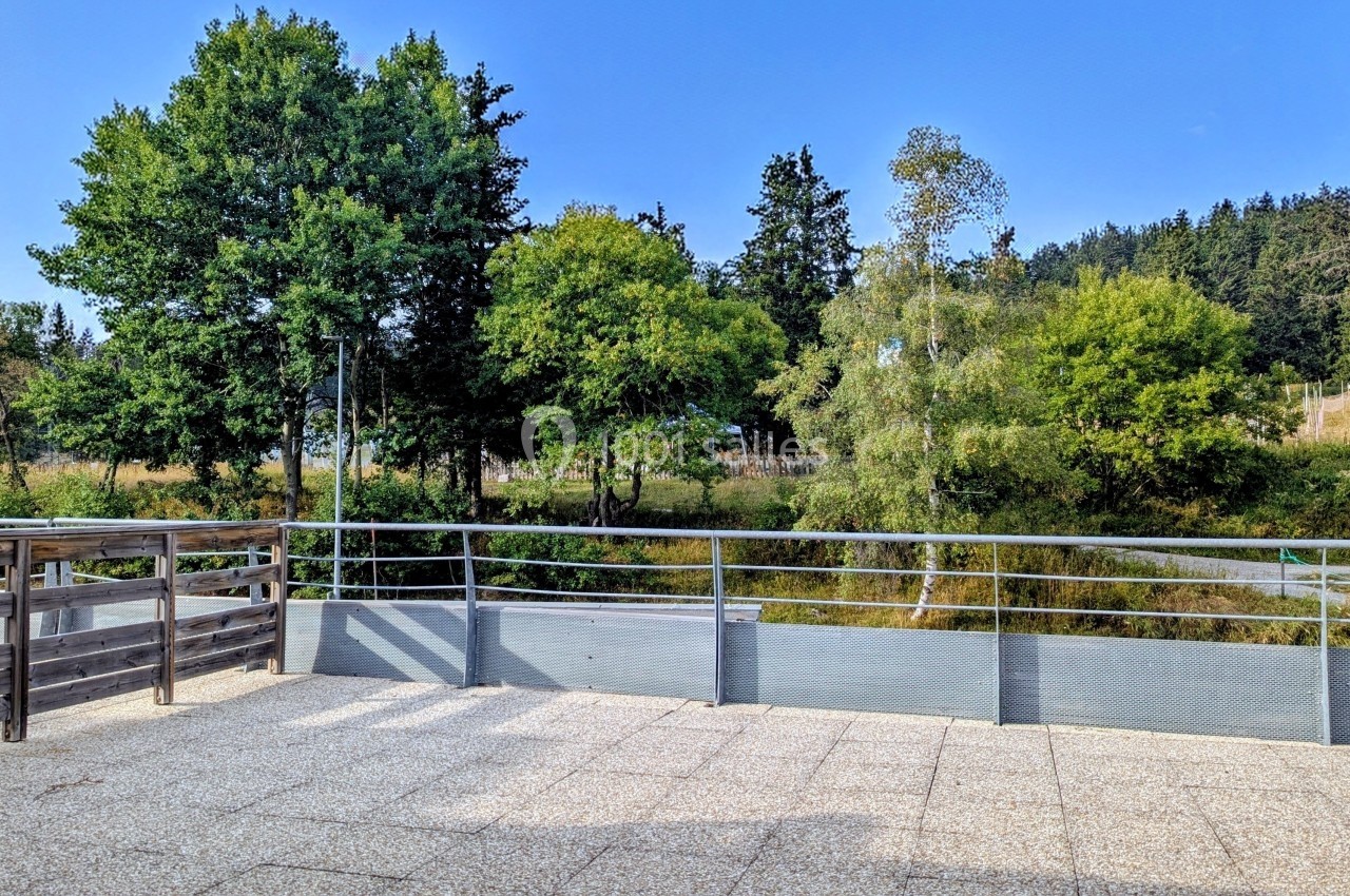 Terrasse en dalles claires avec garde-corps métallique, donnant sur un paysage arboré sous un ciel dégagé.