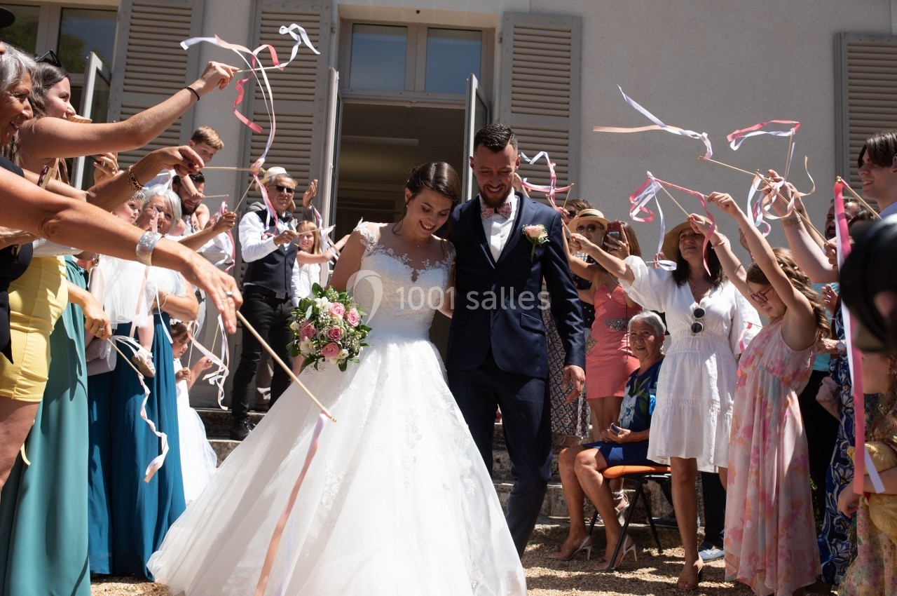 Un couple de mariés souriants sort d'un bâtiment sous des rubans agités par des invités.