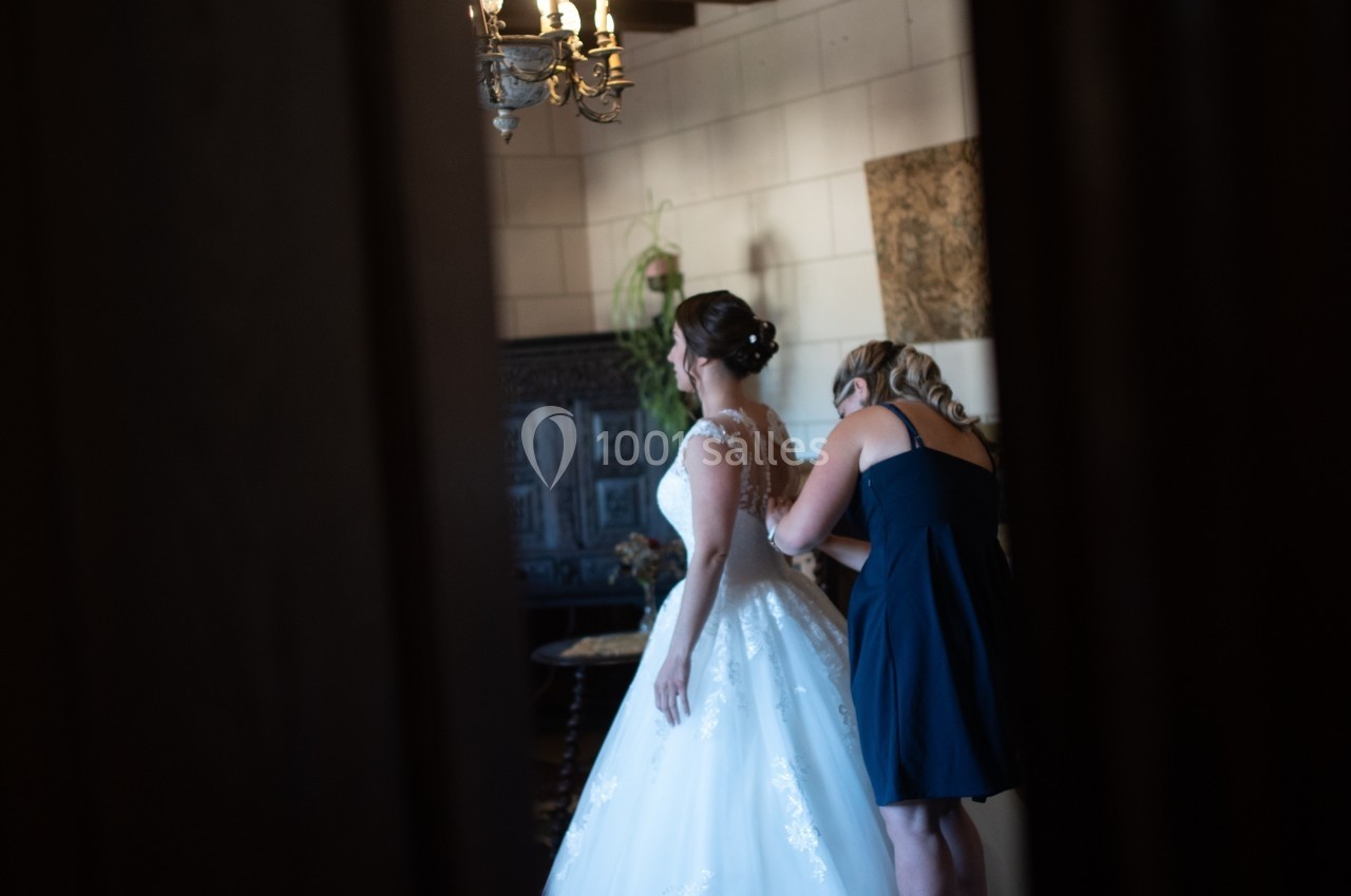 Une femme ajuste la robe de mariée d'une autre dans une pièce éclairée, vue à travers une porte entrouverte.