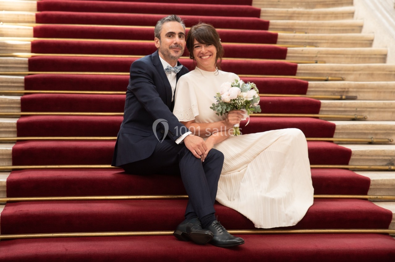 Un couple assis sur des marches recouvertes de tapis rouge, la femme tenant un bouquet de fleurs blanches.