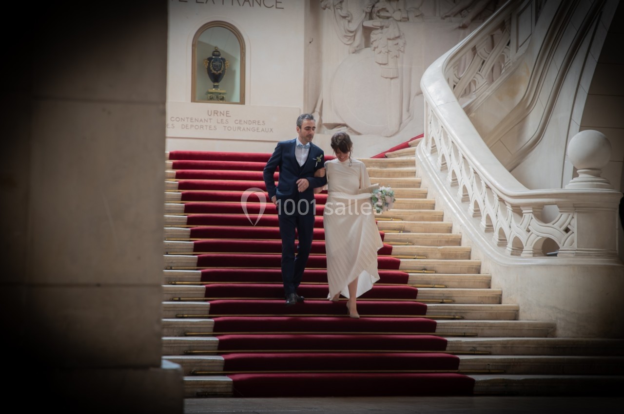 Un couple élégant monte un escalier recouvert d'un tapis rouge dans un bâtiment au décor classique.