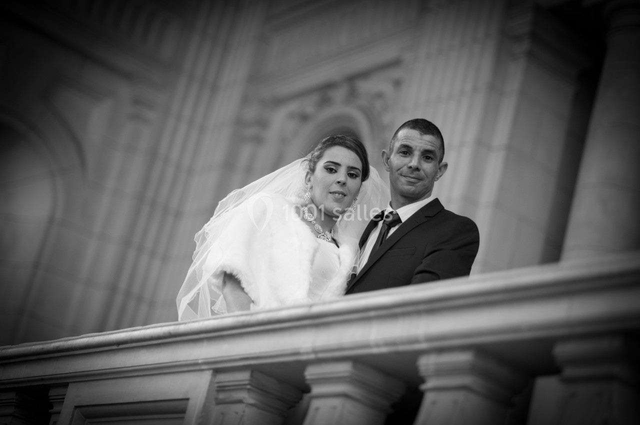 Un couple en tenue de mariage pose sur un balcon en pierre dans un bâtiment à l'architecture classique.