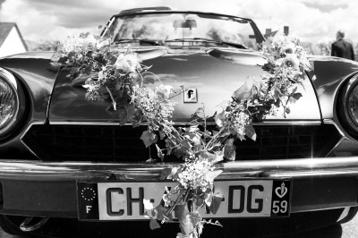 Voiture ancienne décapotable décorée de fleurs pour une cérémonie, stationnée sur une rue pavée.