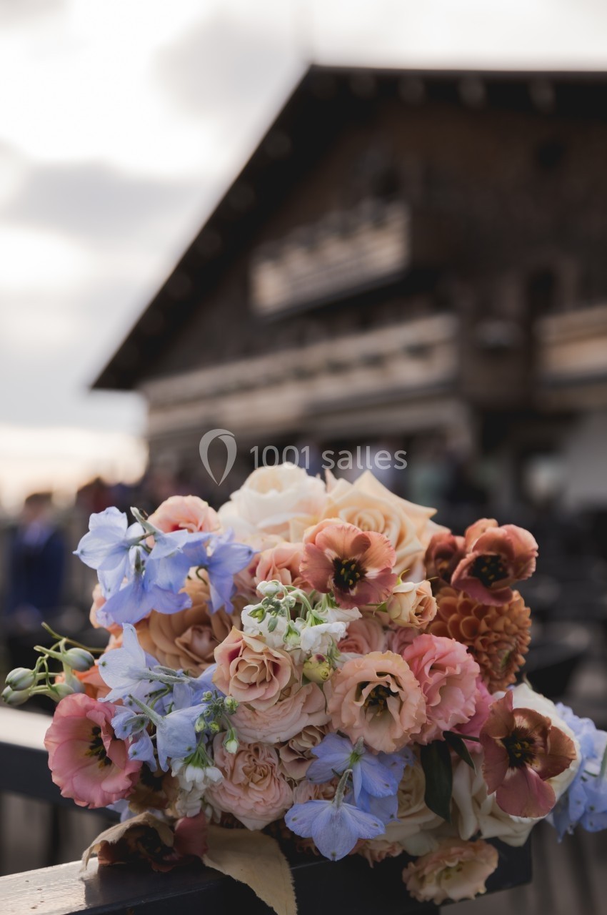 Bouquet de fleurs pastel posé sur une rambarde, avec un bâtiment en bois flou en arrière-plan.