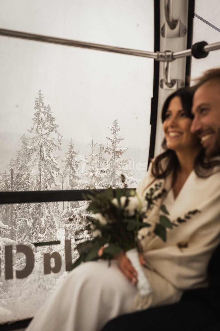 Un couple souriant dans une cabine de téléphérique avec une vue sur une forêt enneigée en arrière-plan.