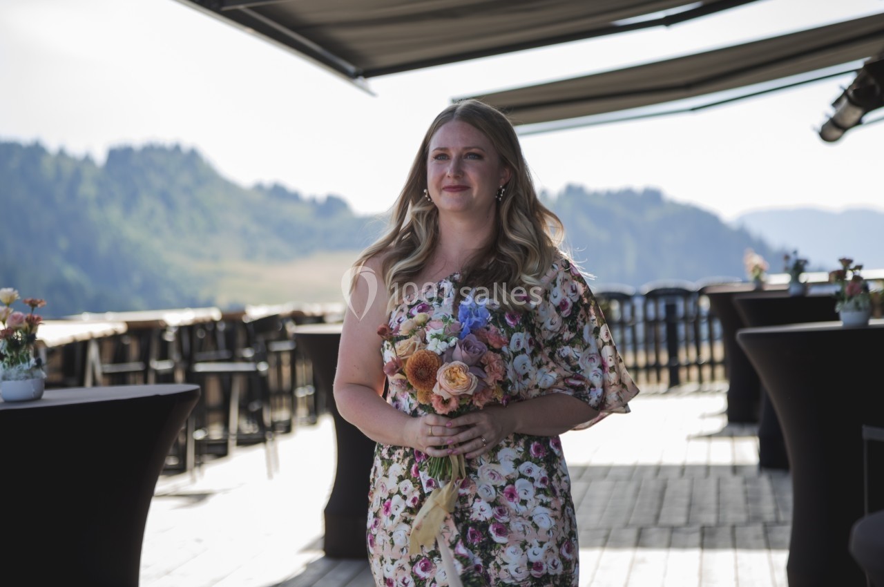 Une femme tenant un bouquet de fleurs marche sur une terrasse en bois avec une vue sur des collines verdoyantes.