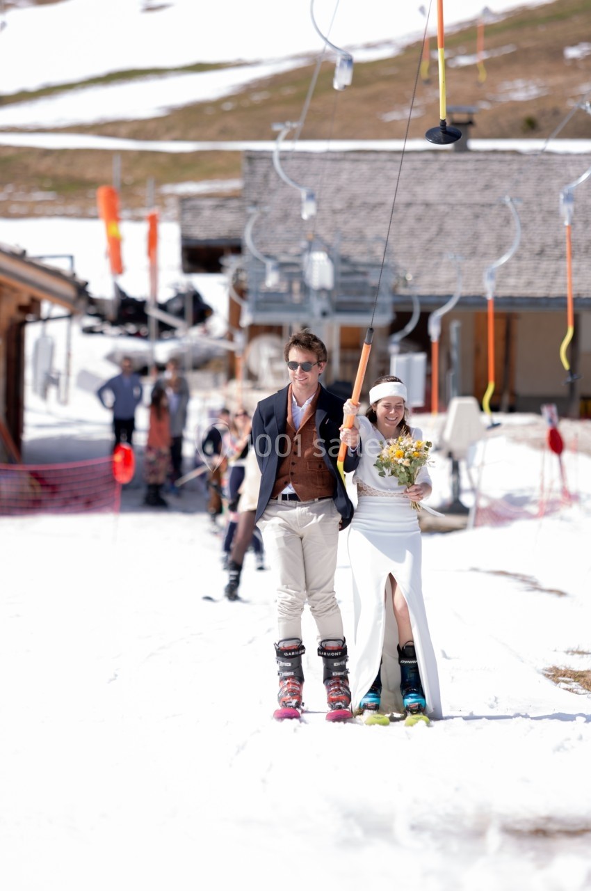 Un couple en tenue de mariage monte un téléski dans une station de ski enneigée par temps ensoleillé.