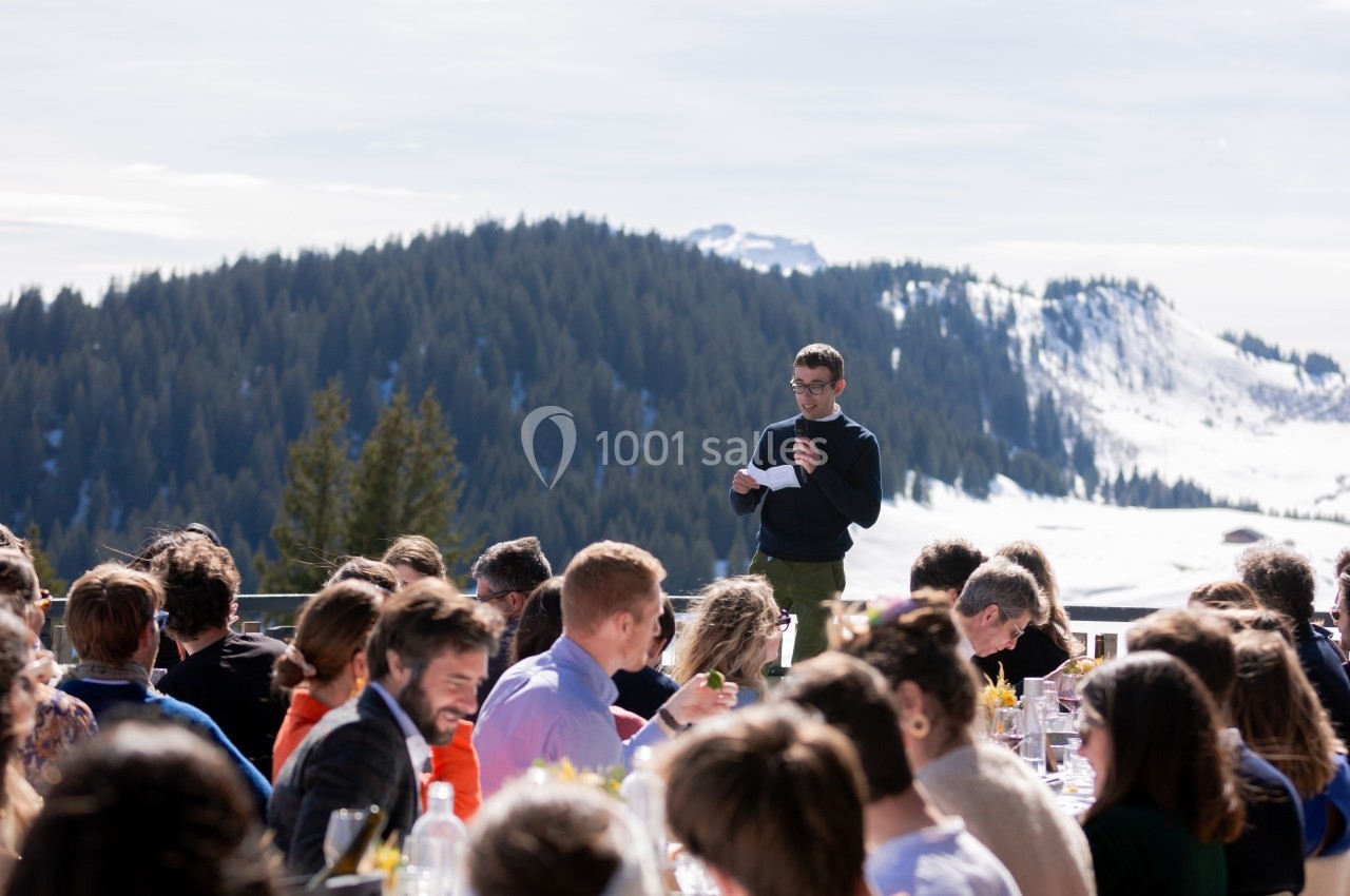 Un homme fait un discours devant un groupe de personnes assises à l'extérieur, avec des montagnes enneigées en arrière-plan.