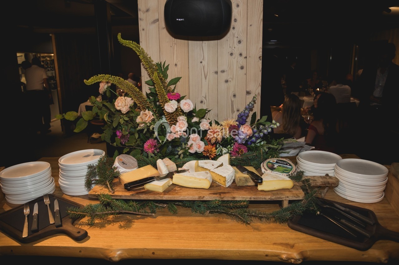 Plateau de fromages variés disposé sur une table en bois, entouré de fleurs et de piles d'assiettes blanches.