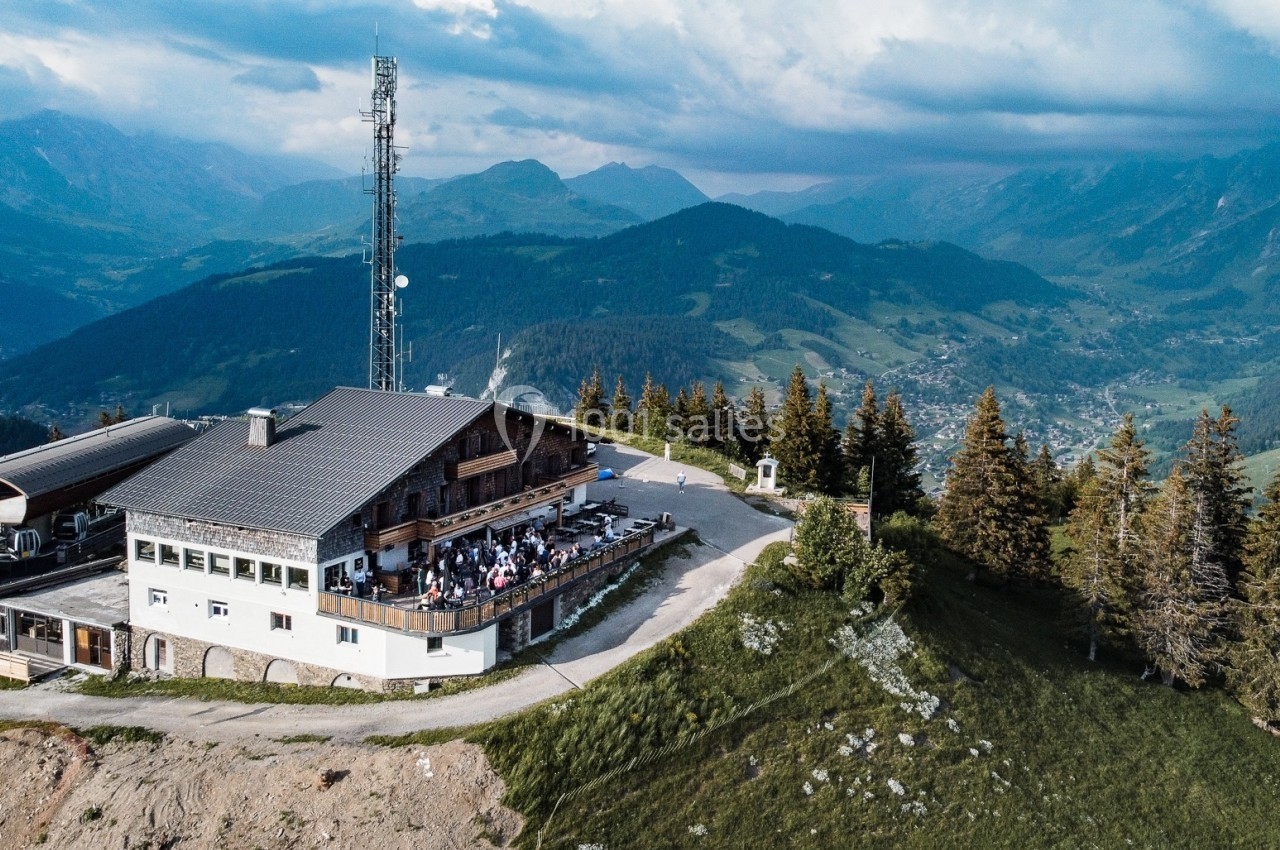 Vue aérienne d'un chalet en montagne avec une terrasse animée, entouré de forêts et de vallées verdoyantes.