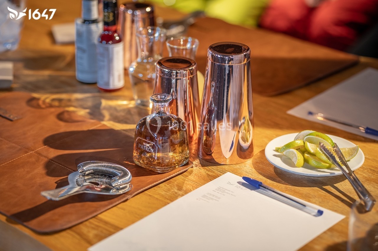 Ustensiles de bar, bouteille de spiritueux, assiette de quartiers de citron et feuilles de papier sur une table en bois.
