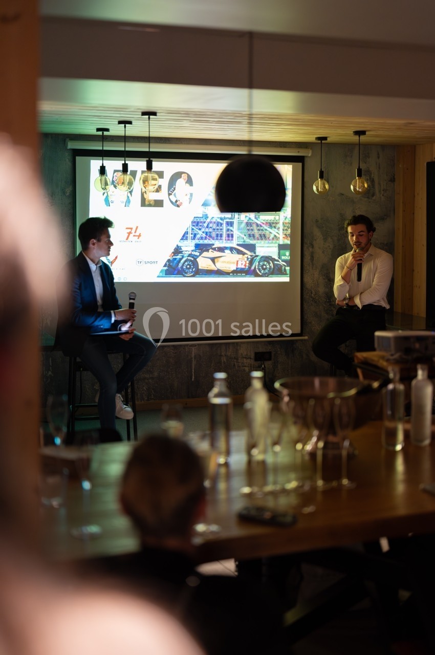 Deux hommes discutent sur une scène devant un écran projetant une image de voiture de course et des portraits.