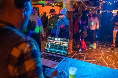 Un DJ mixe sur son ordinateur portable lors d'une soirée animée dans un espace intérieur éclairé par des lumières colorées.