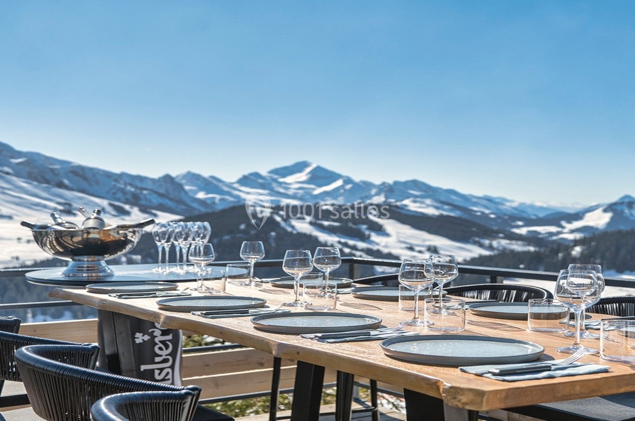 Table en bois dressée avec verres et assiettes, sur une terrasse offrant une vue panoramique sur des montagnes enneigées.