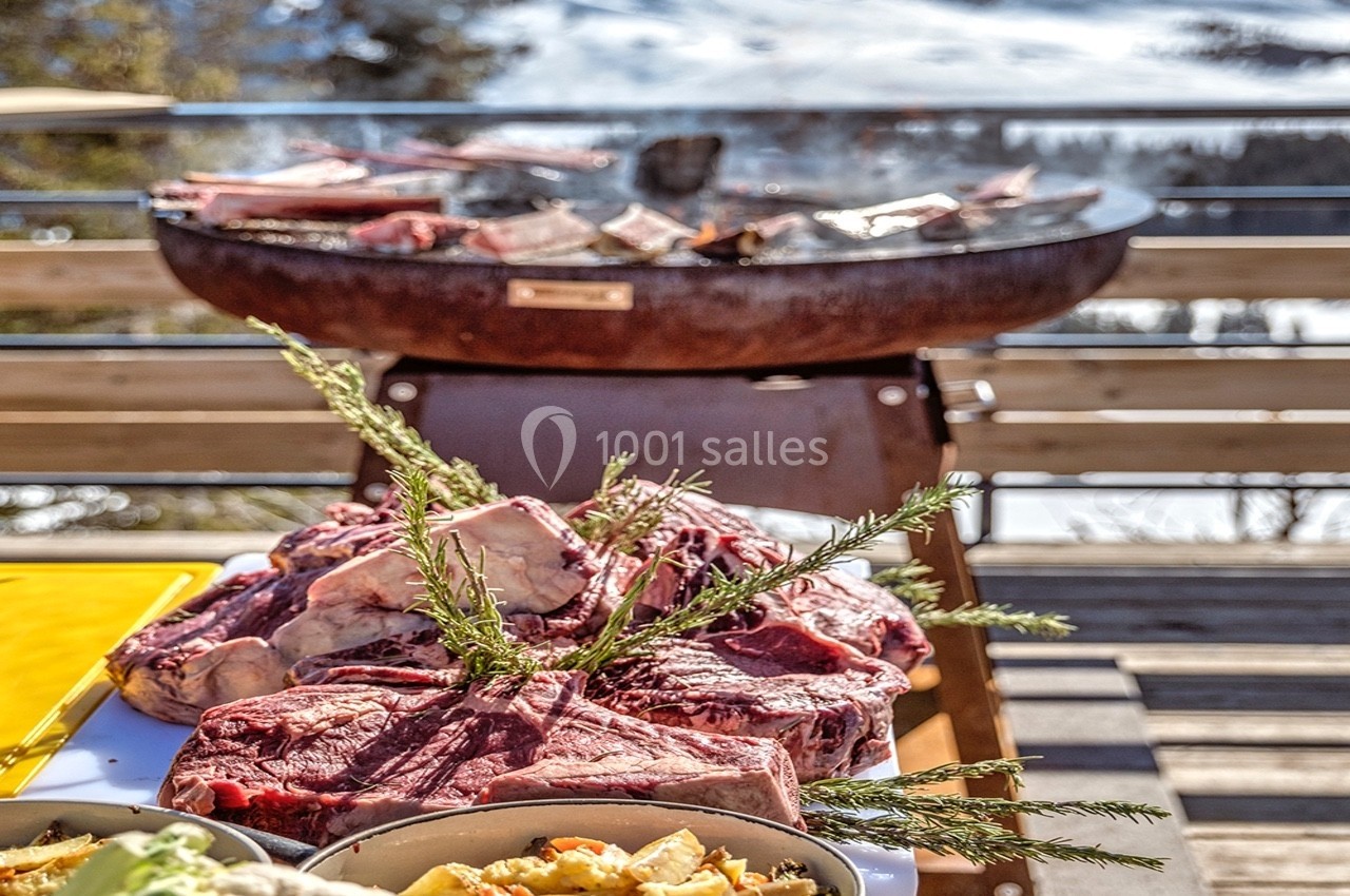 Viande crue et garnitures sur une table devant un barbecue allumé, avec un paysage enneigé en arrière-plan.