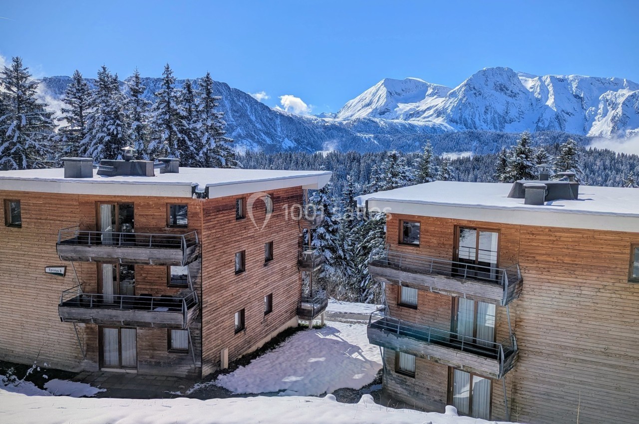Deux bâtiments en bois entourés de neige et de sapins, avec des montagnes enneigées en arrière-plan sous un ciel bleu.