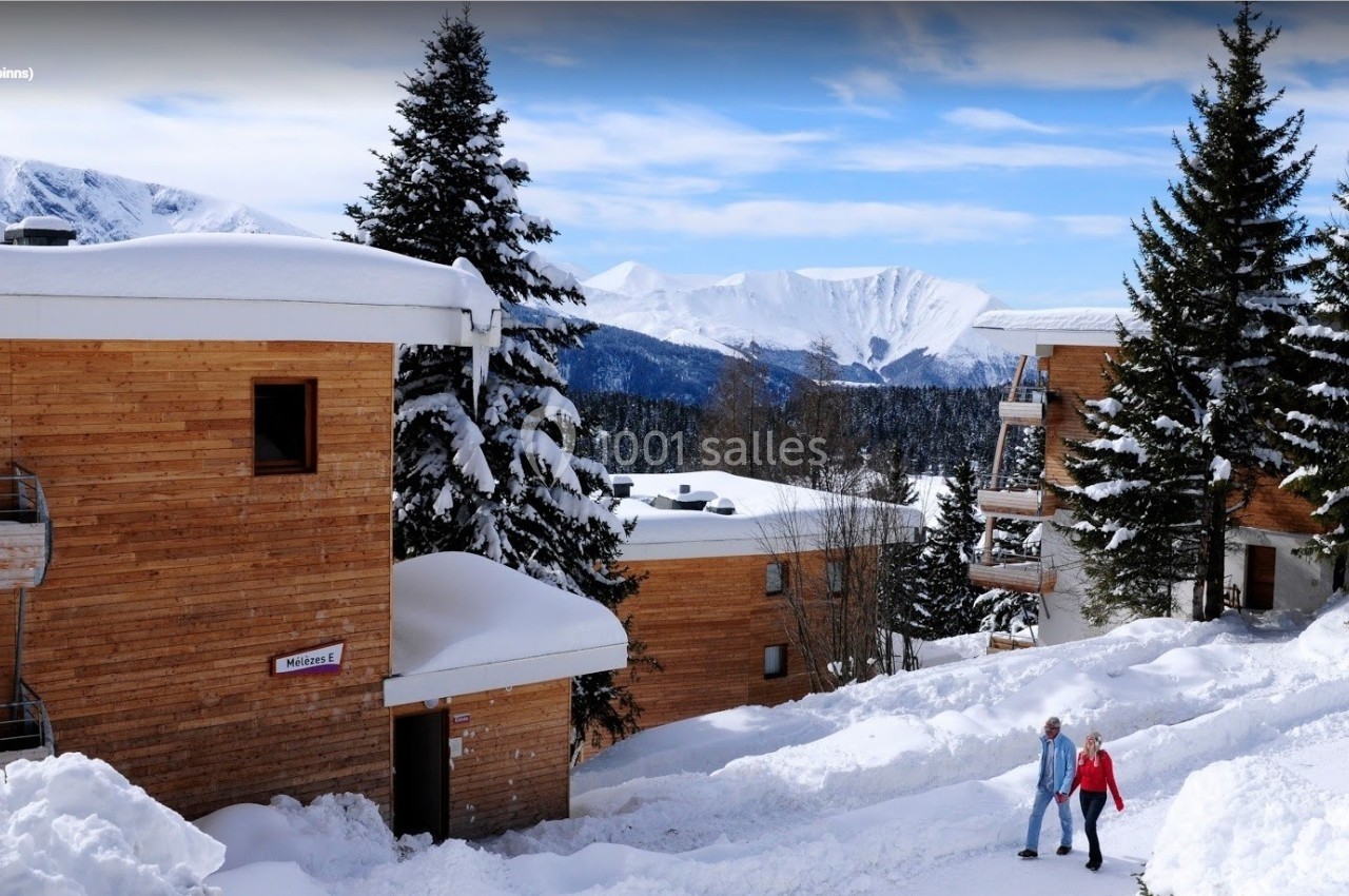 Chalets en bois entourés de neige, avec deux personnes marchant sur un chemin enneigé, montagnes en arrière-plan.