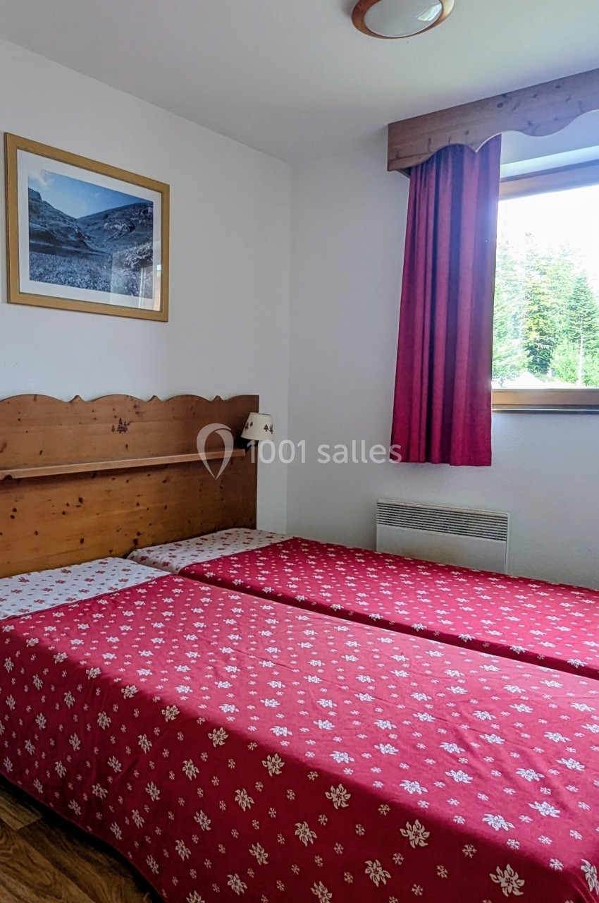 Deux lits simples avec couvre-lits rouges à motifs blancs dans une chambre lumineuse avec fenêtre et décoration en bois.