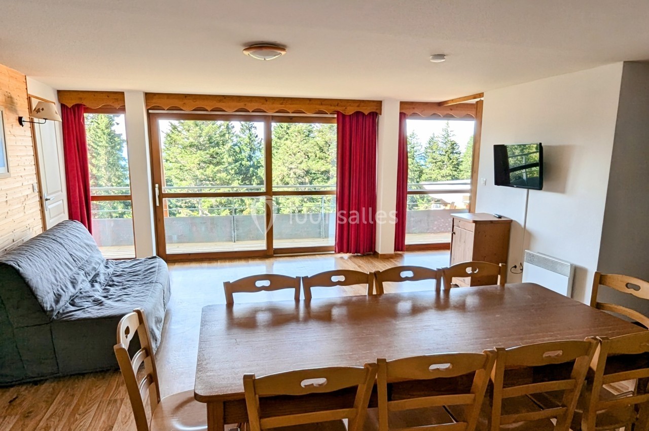 Salle à manger lumineuse avec table en bois, chaises, canapé, télévision murale et grandes baies vitrées donnant sur une…