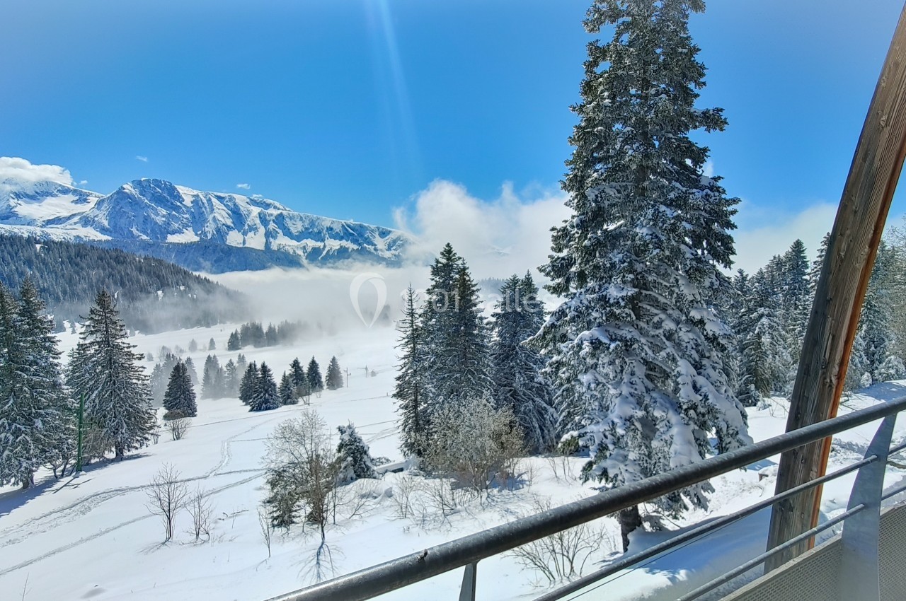 Paysage enneigé avec des sapins, une montagne en arrière-plan et une balustrade au premier plan sous un ciel bleu.