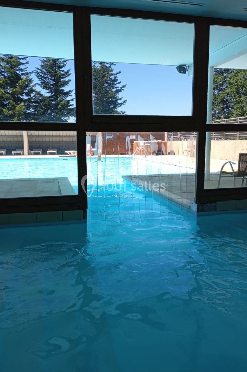 Vue depuis une piscine intérieure donnant sur une piscine extérieure ensoleillée, entourée de chaises longues et d'arbres.