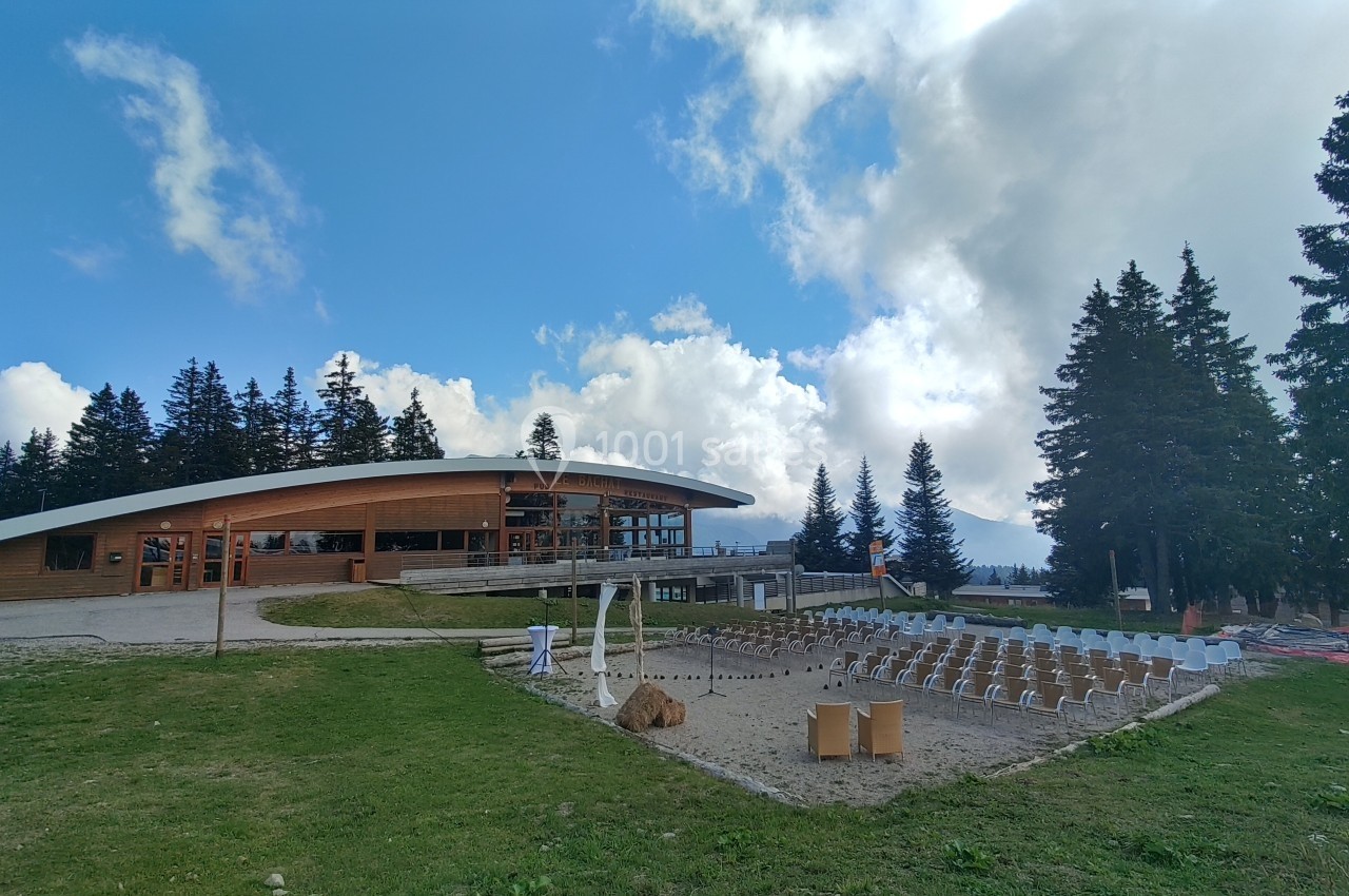 Bâtiment moderne en bois avec terrasse, entouré de sapins et d'une aire aménagée en plein air sous un ciel partiellement…