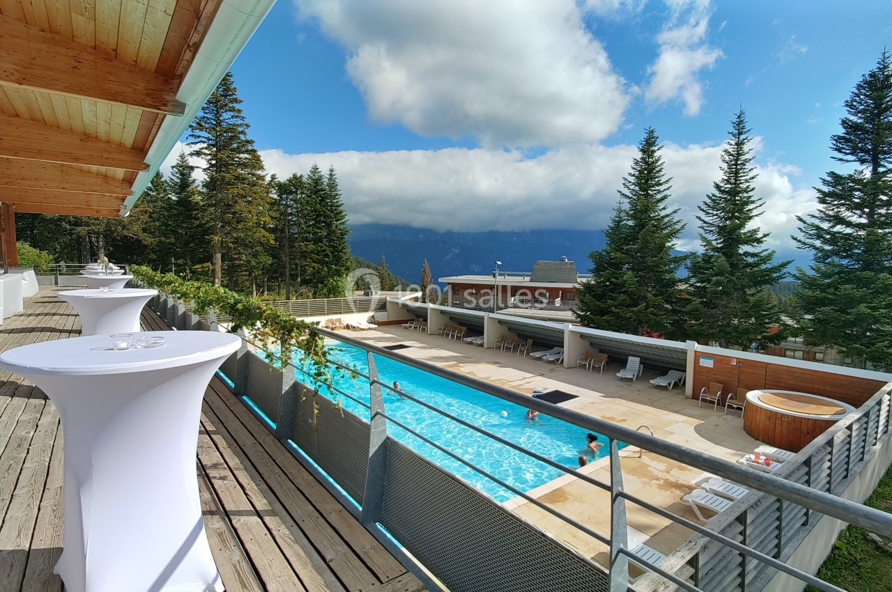 Terrasse en bois surplombant une piscine extérieure entourée de sapins, avec vue sur des montagnes sous un ciel…