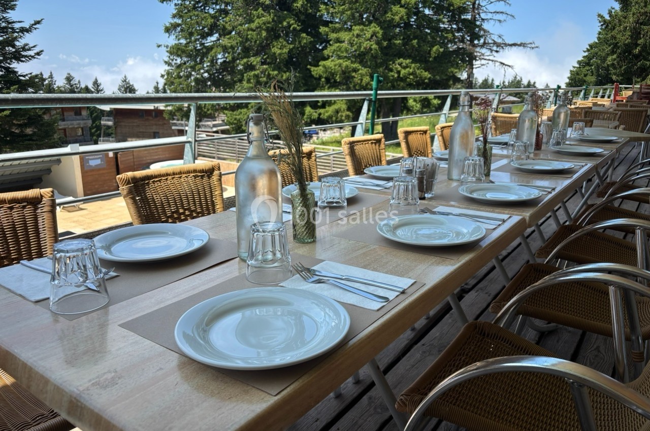 Table en bois dressée en extérieur avec assiettes, verres et carafes, entourée de chaises en osier, vue sur des arbres.