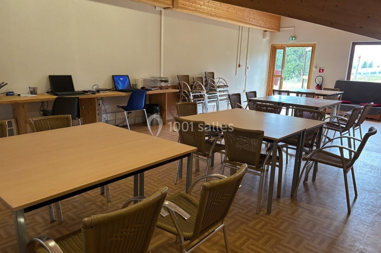 Salle avec des tables en bois, chaises en osier, ordinateurs sur un bureau et une porte donnant sur l'extérieur.