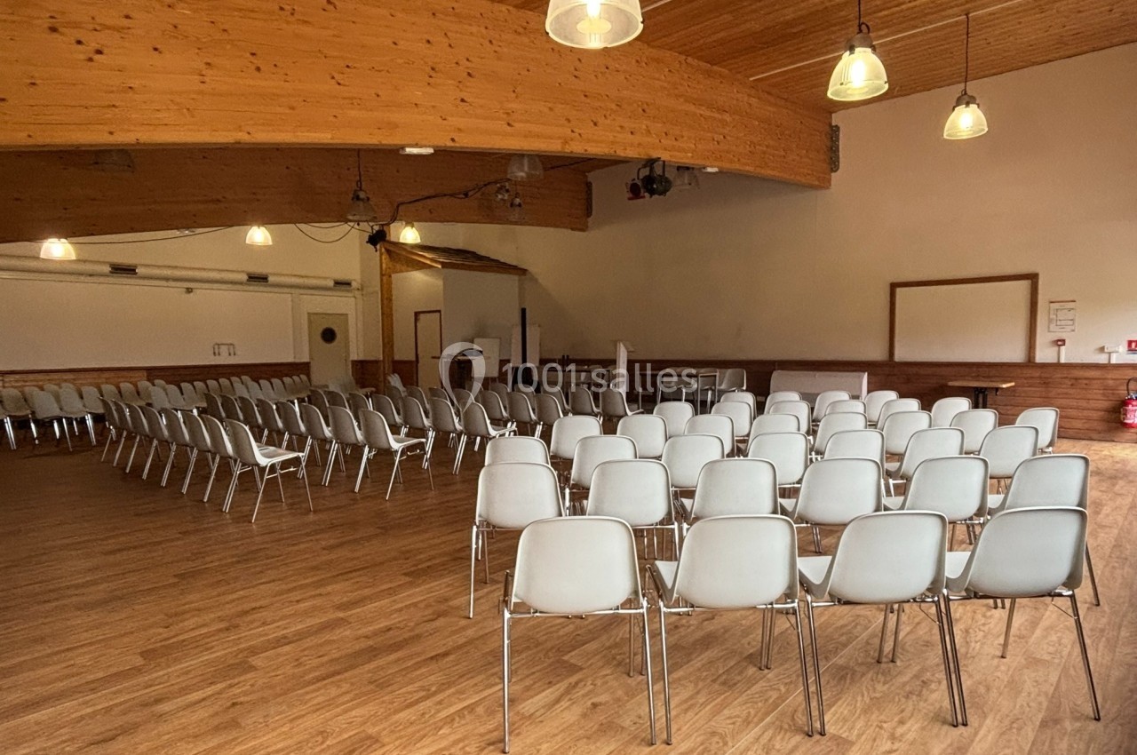 Salle avec parquet, rangées de chaises blanches alignées, éclairage suspendu et estrade en fond.