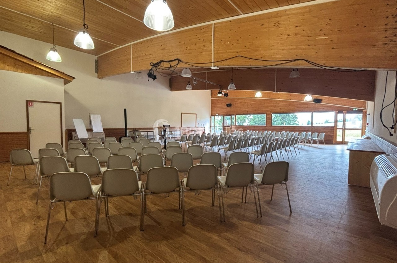 Salle de réunion avec des rangées de chaises alignées, un plafond en bois et de grandes fenêtres laissant entrer la lumière…